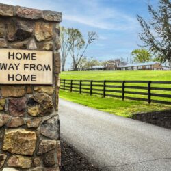 A sign that says "Home Away from Home" is visible on the left side of a horizontal photo. The sign is affixed to a cobblestone-brick pillar that marks the entrance to a road. Paved road leads down a long driveway as the sides show bright green fields and some trees in the distance. Also visible is the Willing Warriors retreat building in the distance. The mulch is freshly placed and the cobble brick is a lot of shades of brown. The sky is a clear with only a few sparse clouds.