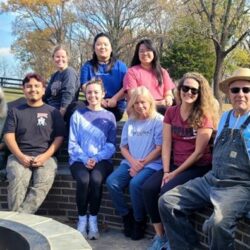 A group of people are posing for a photo while sitting on a brick bench in front of a fire pit. Some are wearing sunglasses and hats, as the setting is outdoors and is in a fenced-in field. Men and women are present, smiling at the camera and wearing casual clothing. A red barn can be seen in the distance, as well as fall foliage.
