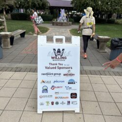 A sign sits on some tiled walkways that eventually lead to a fountain. The sign is white and has a large Willing Warriors logo on it, and a bunch of sponsors for the event, one of which includes BMRA. 2 people can be seen in the background, and a person's peace sign can be seen popping in through the right side of the image. Trees and grass can be seen in the background.