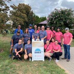 A group of people pose in front of a plastic sign. the side on the left is wearing blue shirts and the side on the right is wearing pink shirts. The group is made of men and women who are smiling, most of which are wearing sunglasses. The setting is outdoors, with lots of grass and trees visible in the background. Some people are kneeling while the taller ones stand.