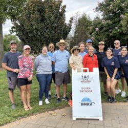 A group of both men and women are posing for a photo around a white sign that says BMRA is a Silver Sponsor of Willing Warriors. There's some people in branded shirts and others that have casual clothing. Some are wearing sunglasses, as this photo was taken outside. There are large, leafy trees in the background and while the people are standing on green grass, the sign is on a tiled walkway. There's a small children's playground in the background and the sky is a little overcast.