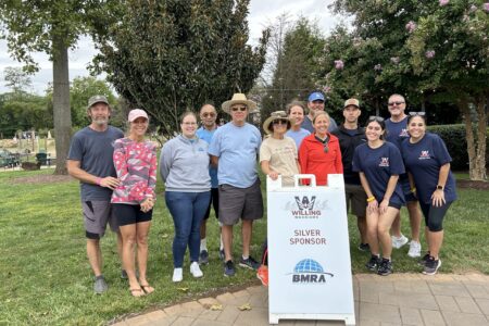 A group of both men and women are posing for a photo around a white sign that says BMRA is a Silver Sponsor of Willing Warriors. There's some people in branded shirts and others that have casual clothing. Some are wearing sunglasses, as this photo was taken outside. There are large, leafy trees in the background and while the people are standing on green grass, the sign is on a tiled walkway. There's a small children's playground in the background and the sky is a little overcast.
