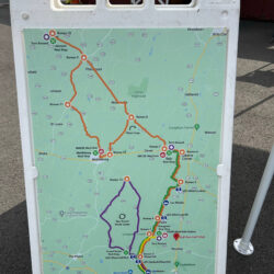 An image of a large plastic floor sign that holds a map with many routes on it. There's one that is the largest in orange, and one that sticks out in purple coloring. The rest of the map is green and looks like a Google Maps image.