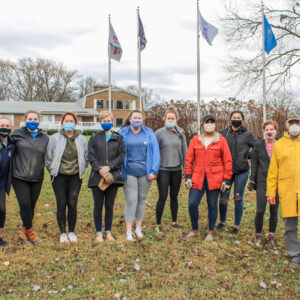 Masked men and women are seen standing in front of six large flagpoles. There's a large residence in the background, enveloped in winter trees with sparse leaves. Fall foliage lies underneath the people's feet, and an overcast sky looms overhead. Everyone is dressed casually and wearing masks.