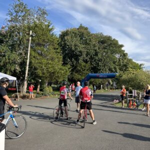 A lot of people are gathered at the beginning of a bike race. There's a white tent on the left side, and a few bikers in the middle of the frame. Some bystanders watch on as the bikers prepare for the start. A large blue, inflatable banner sits between trees in the background and signify the starting area. A blue sky with some wispy clouds can be seen overhead. Pink blossoms can be seen on some of the trees.