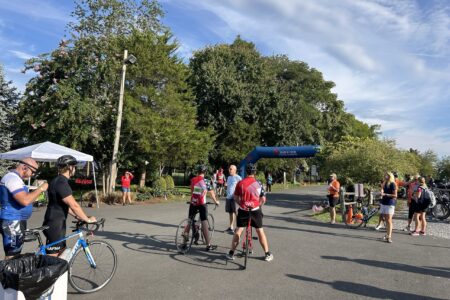 A lot of people are gathered at the beginning of a bike race. There's a white tent on the left side, and a few bikers in the middle of the frame. Some bystanders watch on as the bikers prepare for the start. A large blue, inflatable banner sits between trees in the background and signify the starting area. A blue sky with some wispy clouds can be seen overhead. Pink blossoms can be seen on some of the trees.