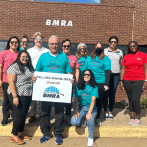 A group of men and women stand in front of the BMRA business location, wearing BMRA-Willing-Warrior attire. Most are wearing sunglasses and there's one person in the front holding a sign that says BMRA is a sponsor of Willing Warriors. The group is made of smiling people, and some are knelt down in front of the group. BMRA in the community.