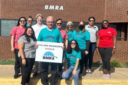 A group of men and women stand in front of the BMRA business location, wearing BMRA-Willing-Warrior attire. Most are wearing sunglasses and there's one person in the front holding a sign that says BMRA is a sponsor of Willing Warriors. The group is made of smiling people, and some are knelt down in front of the group. BMRA in the community.