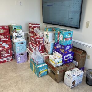 A large pile of diapers sits in the corner of the foyer of the BMRA office building. On the right wall, there's a television mounted that displays a calendar. The boxes of diapers and wipes stands four or five high in places, and there are many brand names like Pampers, Luvs, Huggies, and Kirkland.