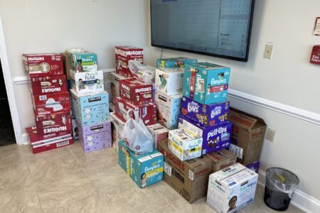A large pile of diapers sits in the corner of the foyer of the BMRA office building. On the right wall, there's a television mounted that displays a calendar. The boxes of diapers and wipes stands four or five high in places, and there are many brand names like Pampers, Luvs, Huggies, and Kirkland.