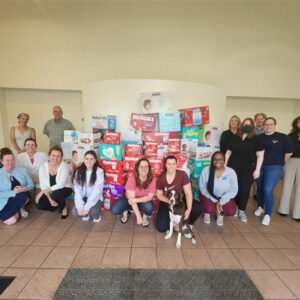 A group of people are posing for a photo in front of a large collection of donated baby supplies, including diapers and wipes from brands like Huggies, Pampers, and BabyGanics. The group consists of both men and women, some standing and some kneeling in front of the diaper donation pile. There is also a dog sitting in front with one person. The setting is indoors with tiled flooring and beige walls. BMRA in the community.
