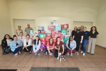 A group of people are posing for a photo in front of a large collection of donated baby supplies, including diapers and wipes from brands like Huggies, Pampers, and BabyGanics. The group consists of both men and women, some standing and some kneeling in front of the diaper donation pile. There is also a dog sitting in front with one person. The setting is indoors with tiled flooring and beige walls. BMRA in the community.