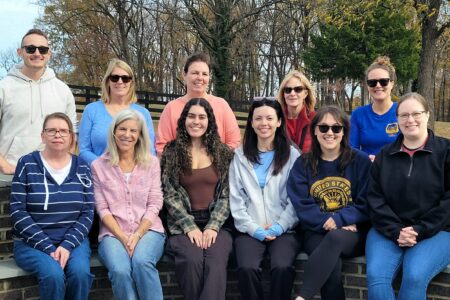 The staff members of BMRA pose for a picture together outside on the Willing Warriors Retreat estate. Half the members are sitting on a brick and cement bench, with the other half of the members standing behind them. They are wearing clothes suited for autumn weather and some are wearing sunglasses.